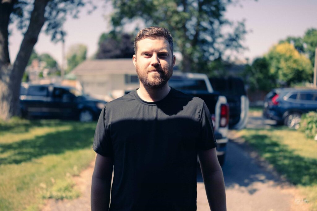 Portrait of a bearded man in a black shirt standing outdoors, with sunlight and shadows across his face, captured in a candid street-style photograph